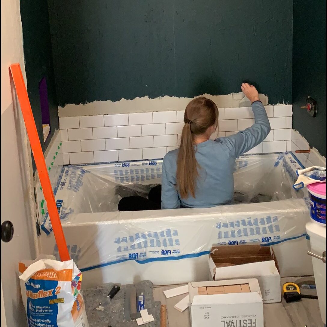 Woman tiling shower wall above bathtub using white subway tile